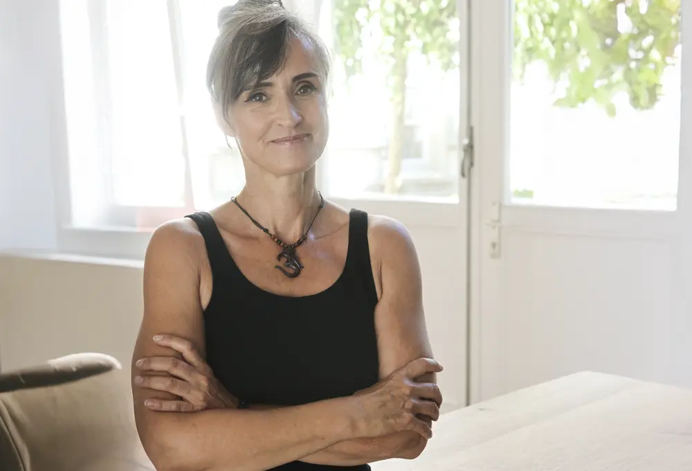 Eine lächelnde Frau steht mit verschränkten Armen vor einem hellen Hintergrund. Sie trägt ein schwarzes Oberteil und eine Halskette mit einem dekorativen Anhänger. Ihr Haar ist zu einem Dutt frisiert, und ihre Ausstrahlung vermittelt Selbstbewusstsein und Freundlichkeit. Durch die Fenster im Hintergrund strömt natürliches Licht in den Raum.